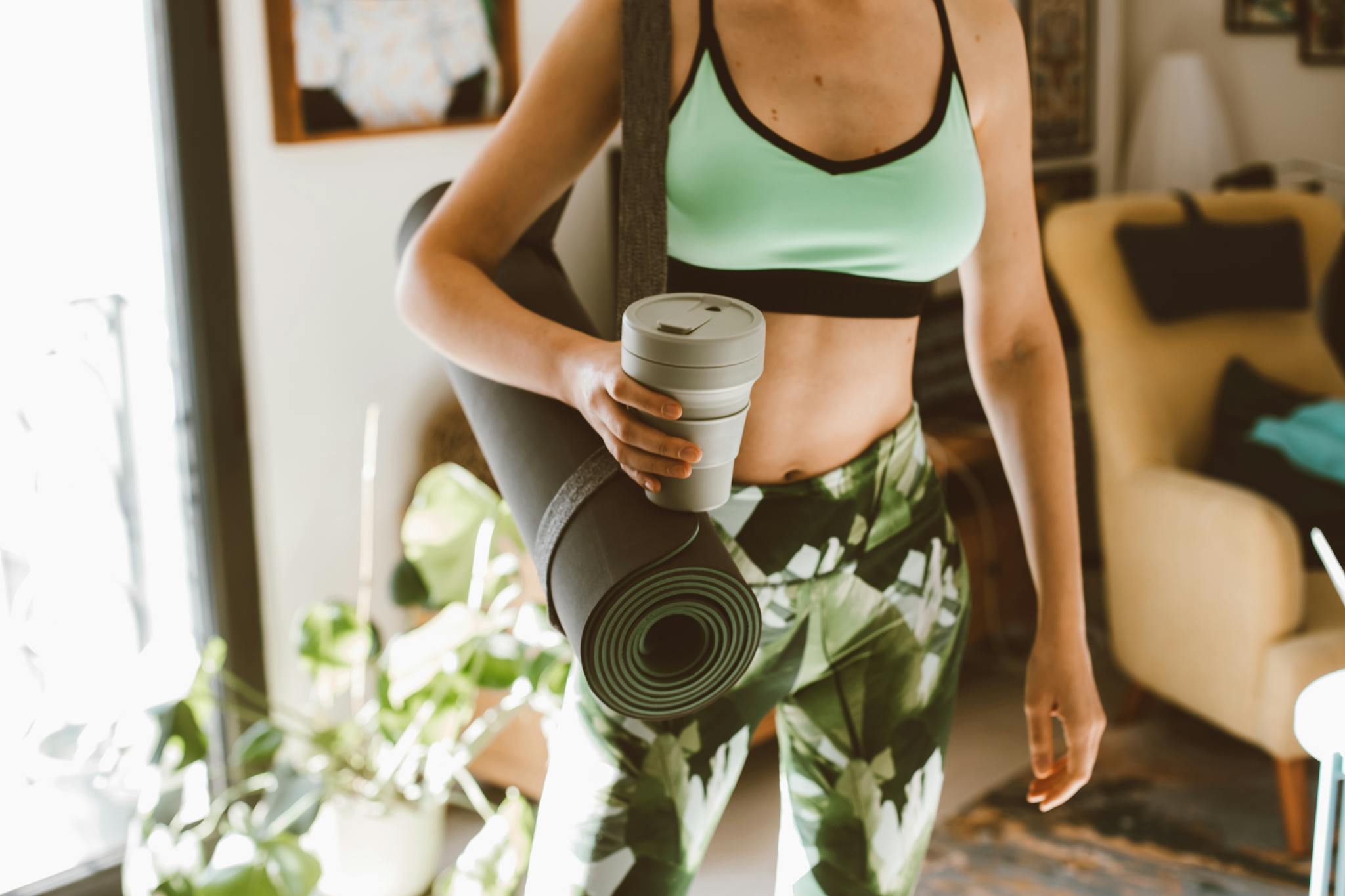 Woman in activewear holding a yoga mat and reusable cup indoors, promoting eco-friendly lifestyle.